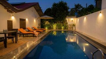 A swimming pool with lounge chairs and a table, surrounded by walls and vegetation.