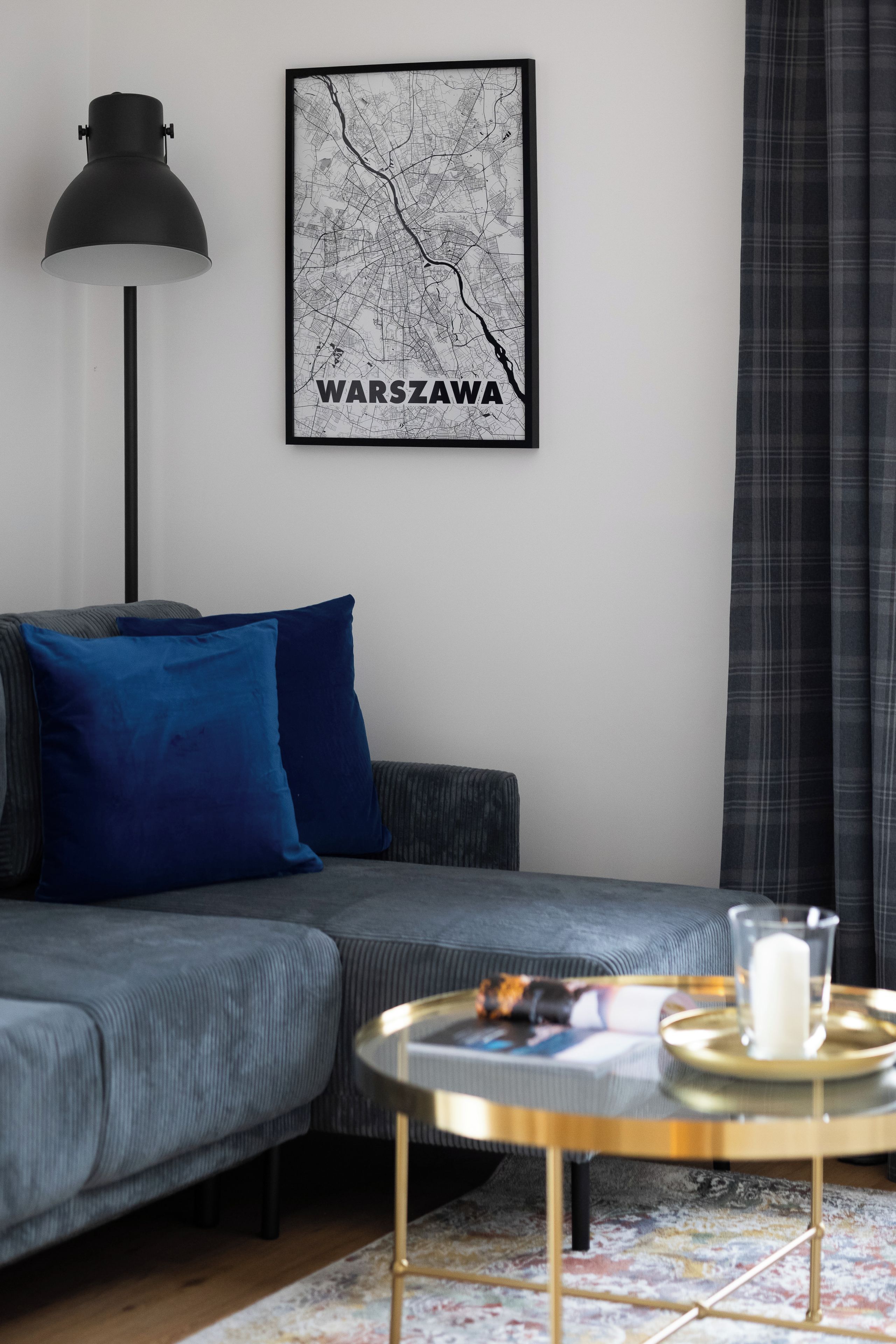 Una sala moderna con un sofá, un mapa enmarcado de Varsovia, una lámpara de pie y una mesita de centro con una vela.