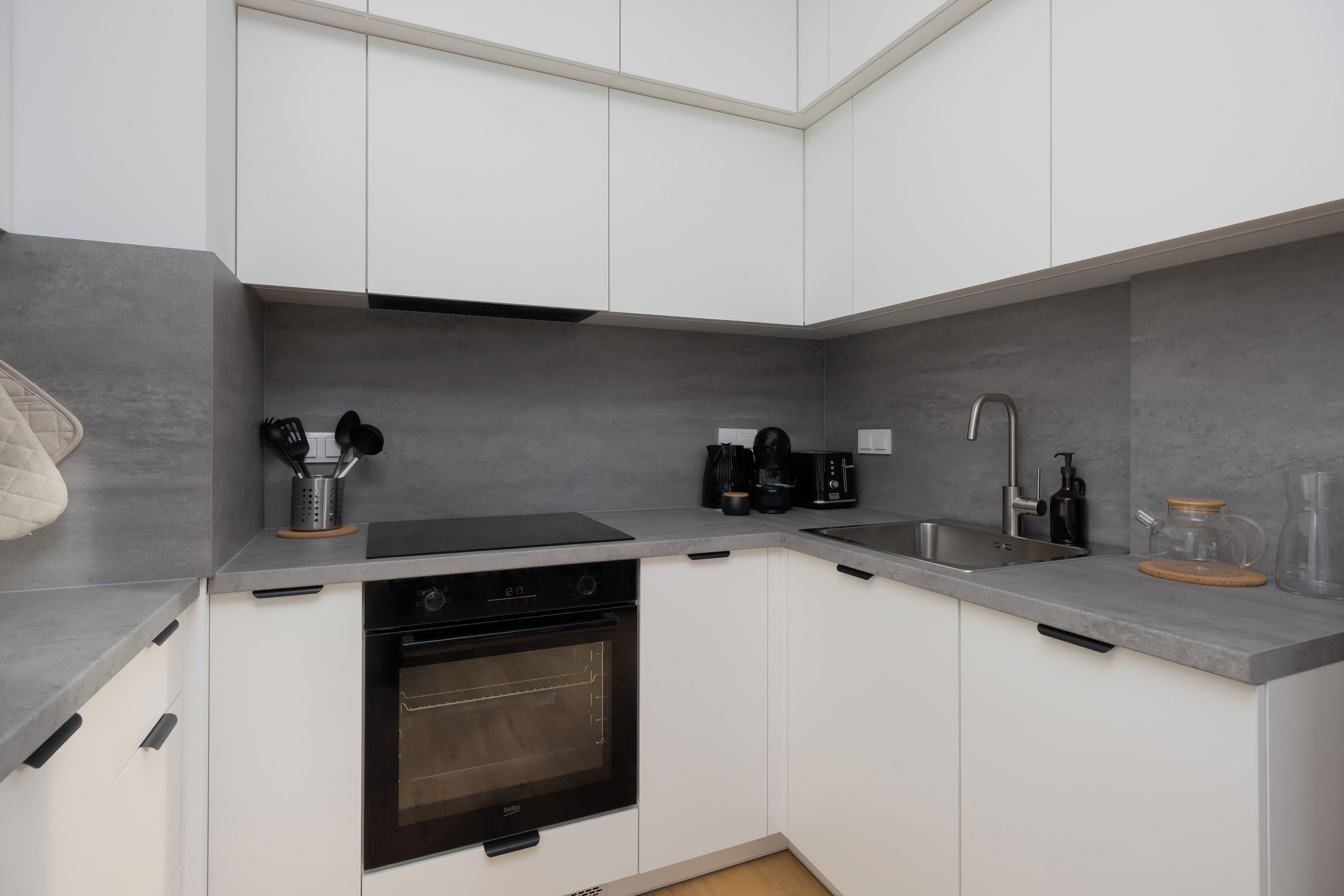 Una cocina compacta con armarios blancos, horno empotrado, cafetera y lavamanos.
