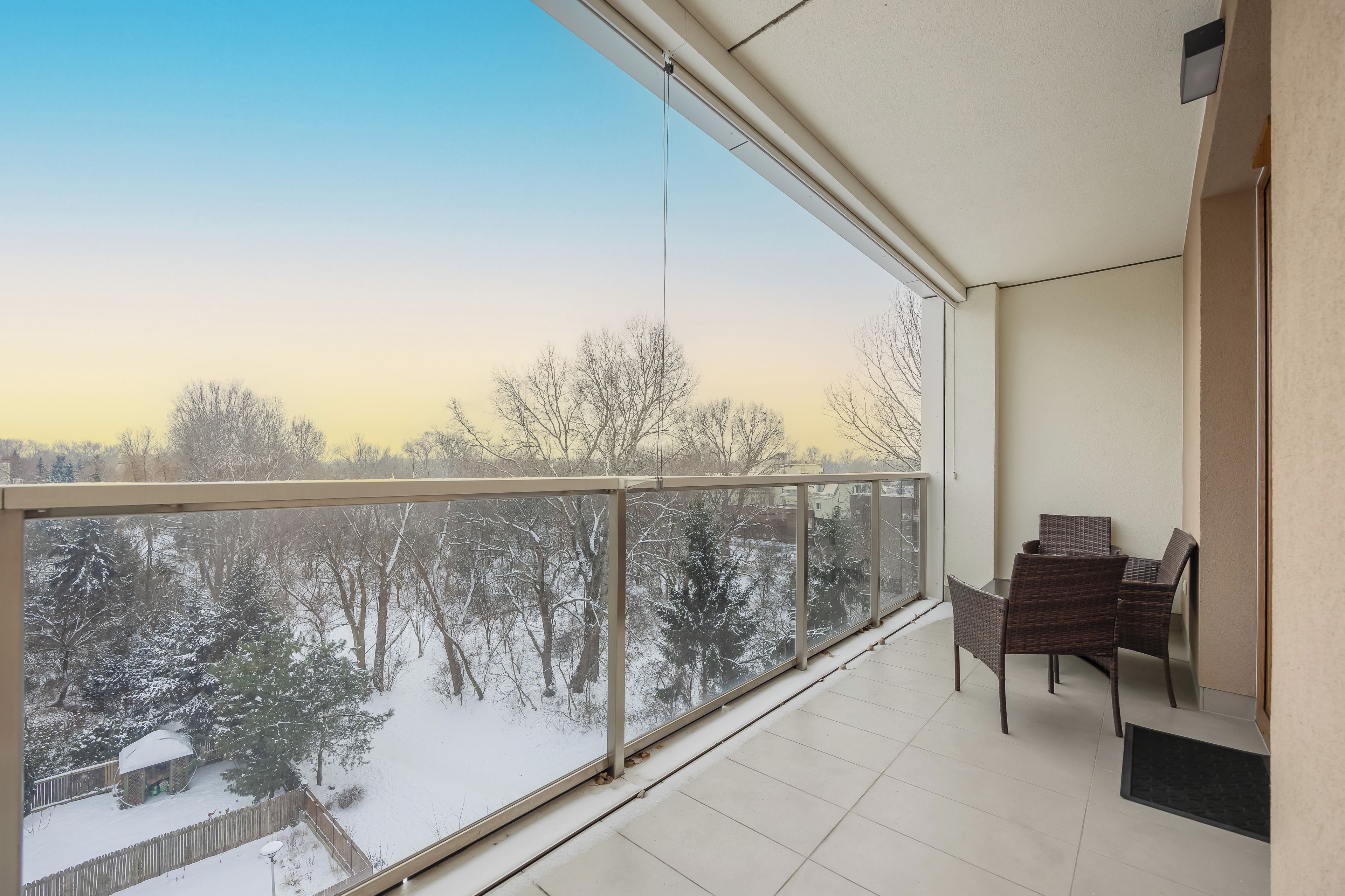 Un balcón con vistas a un paisaje nevado, barandilla de cristal y zona de estar con sillas.