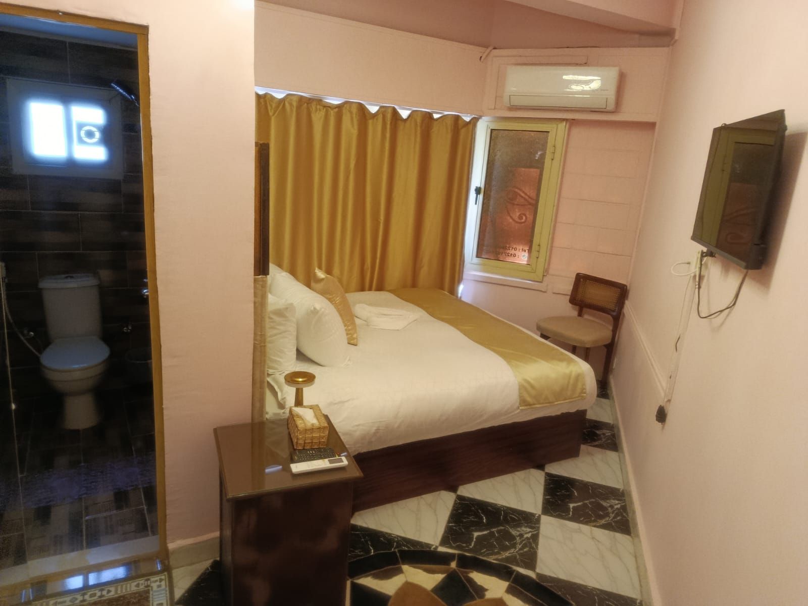 A hotel room with a bed, a bedside table with a lamp, a chair, a small table, a mirror, and an air conditioning unit.