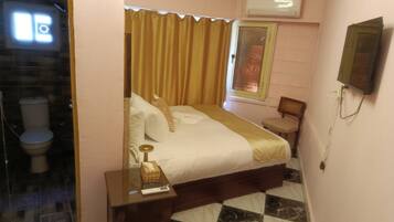 A hotel room with a bed, a bedside table with a lamp, a chair, a small table, a mirror, and an air conditioning unit.