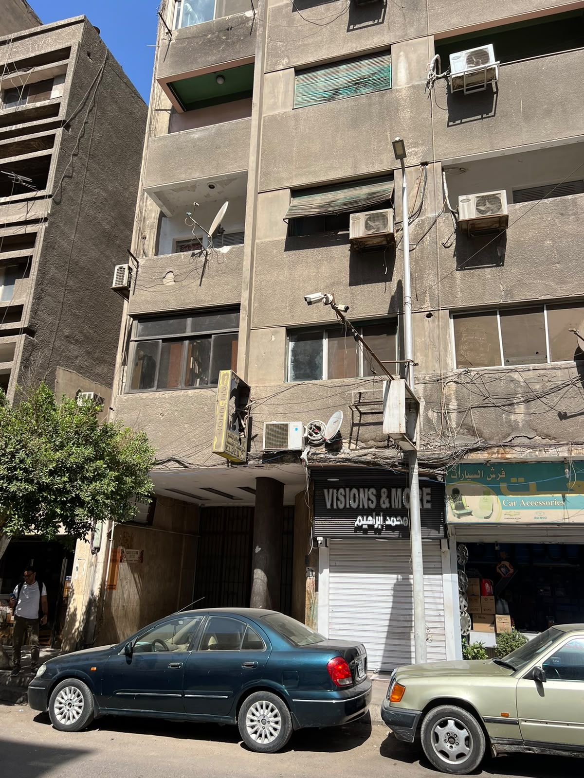 A multi-story building with several air conditioning units, a storefront with a sign, and parked cars.