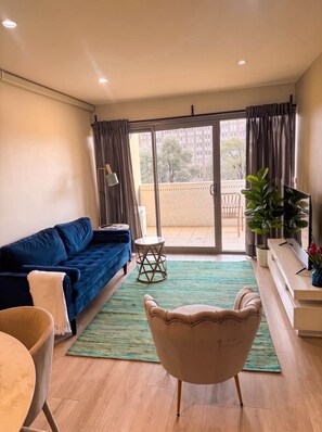 A living room with a blue sofa, a small round table, a chair, a green rug, and a balcony with a view of buildings. - Braddon Luxe Retreat ,Parking, Gym & City Views (Braddon)