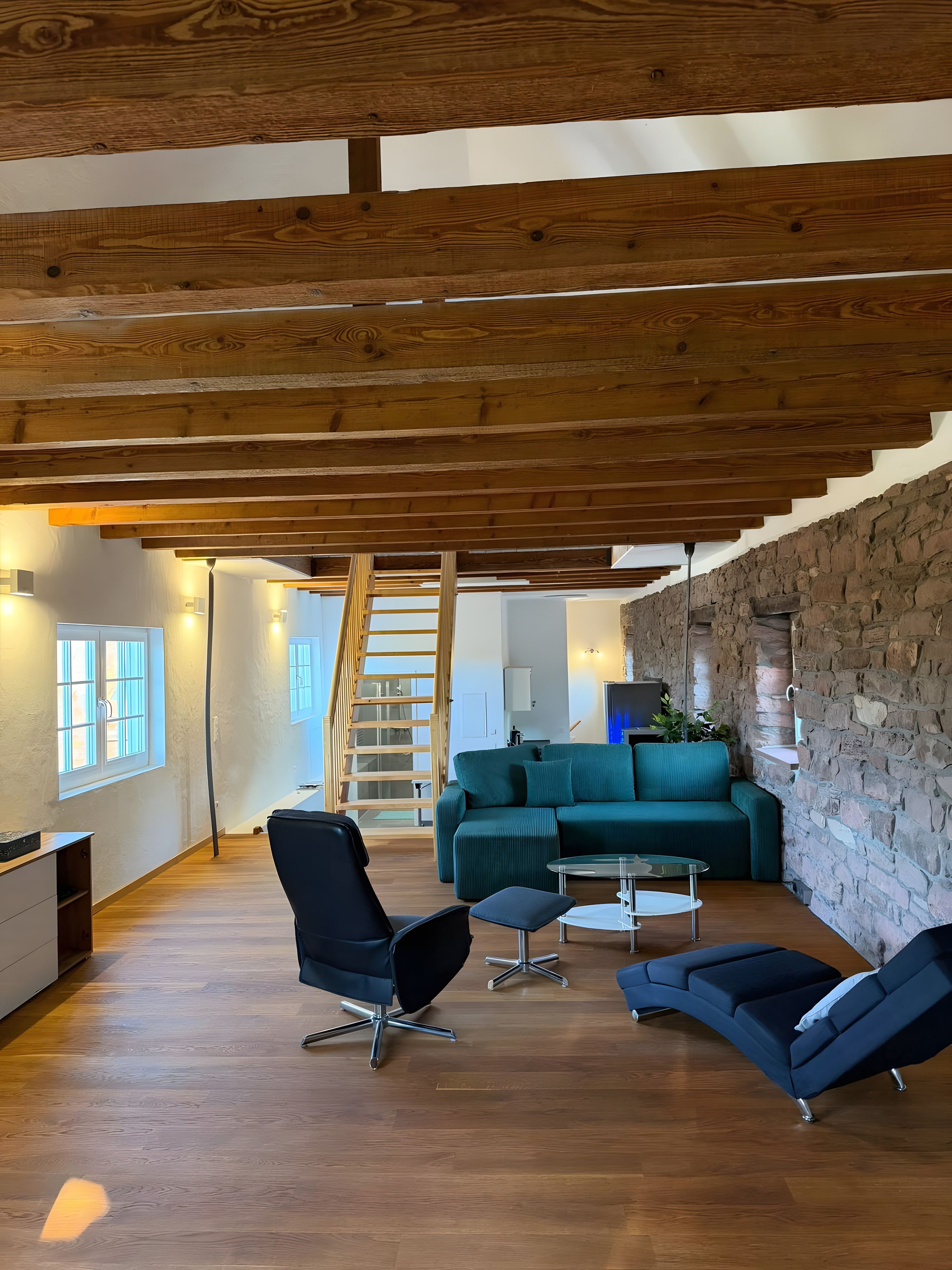A modern living room with a wooden ceiling, a stone wall, and a staircase.