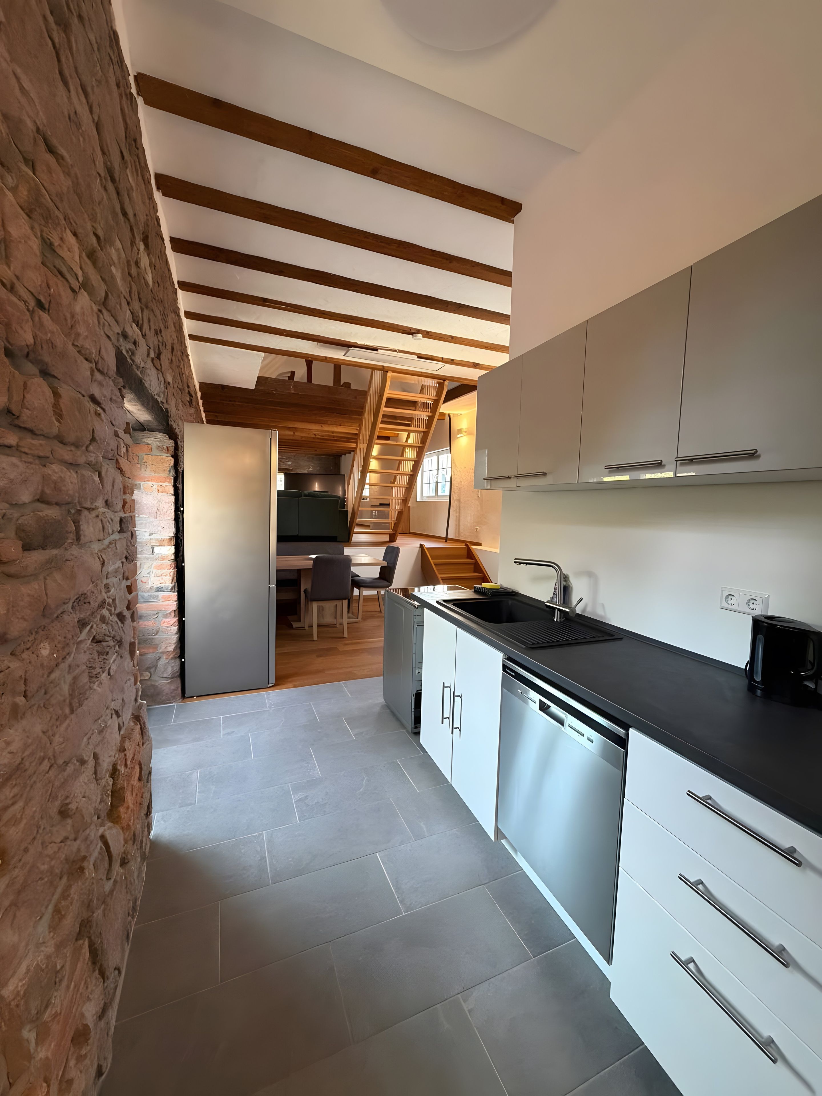 A modern kitchen with a stone wall, wooden beams, and a staircase leading to an upper level.