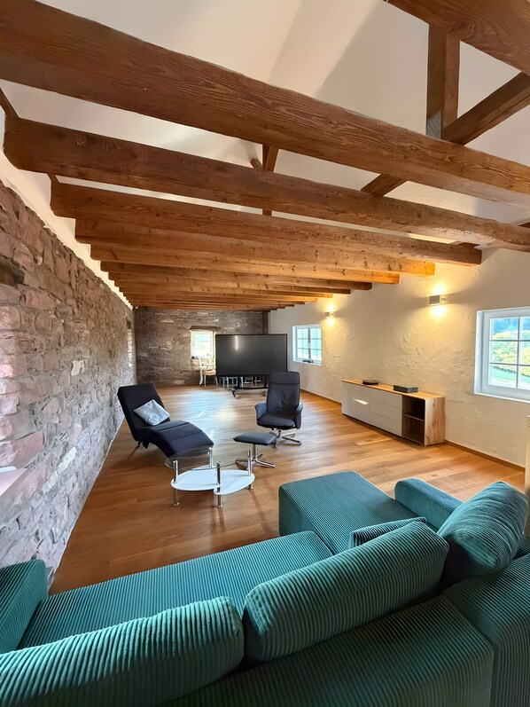 Um amplo loft com vigas de madeira aparentes, uma parede de pedra, um sofá grande e uma TV ao fundo.