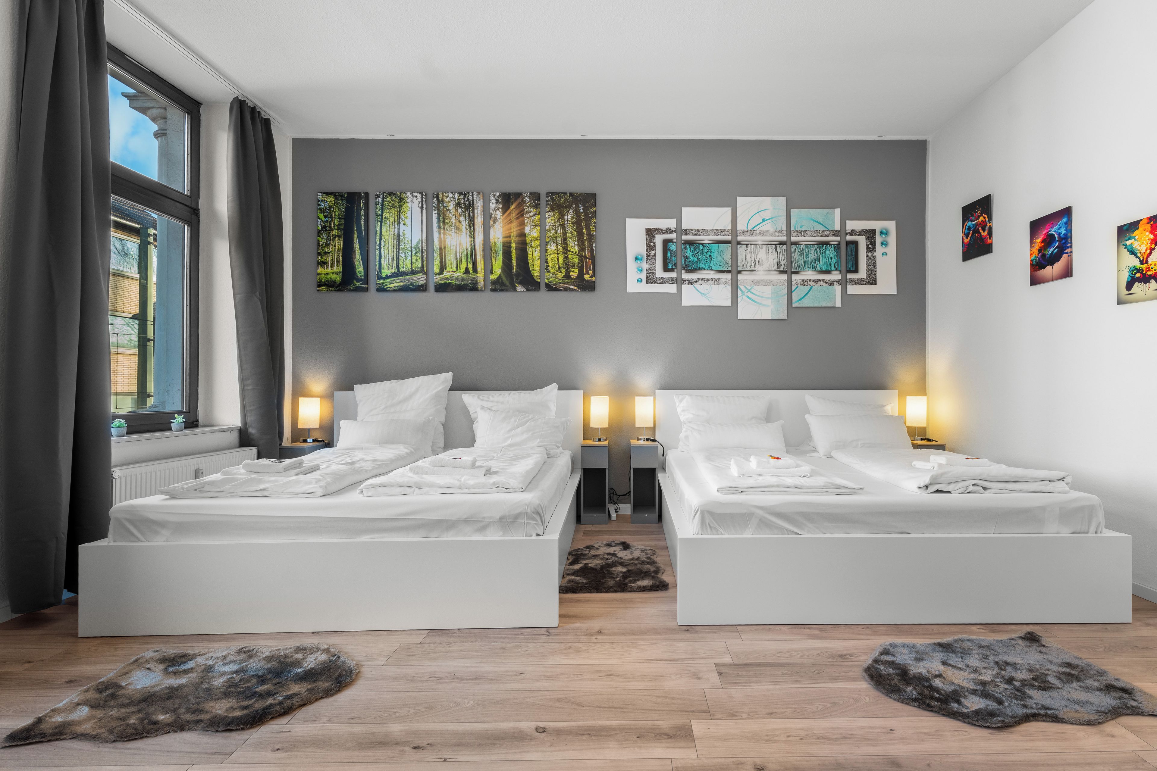 A hotel room with two beds, a large window with curtains, a wall with multiple artworks, and a wooden floor.