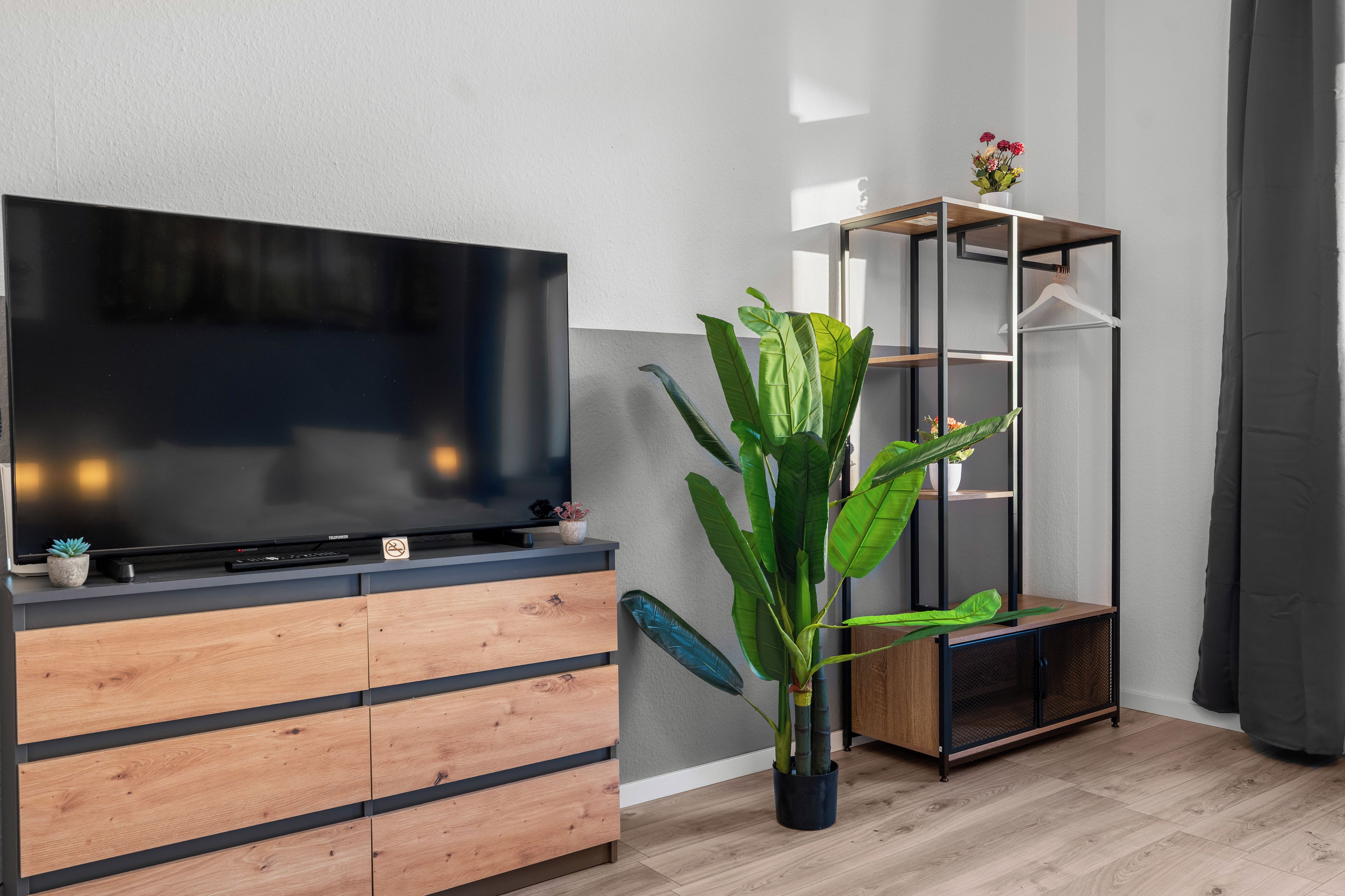 Una sala de estar moderna con un televisor de pantalla plana sobre un mueble de madera, una planta verde grande y una estantería metálica.
