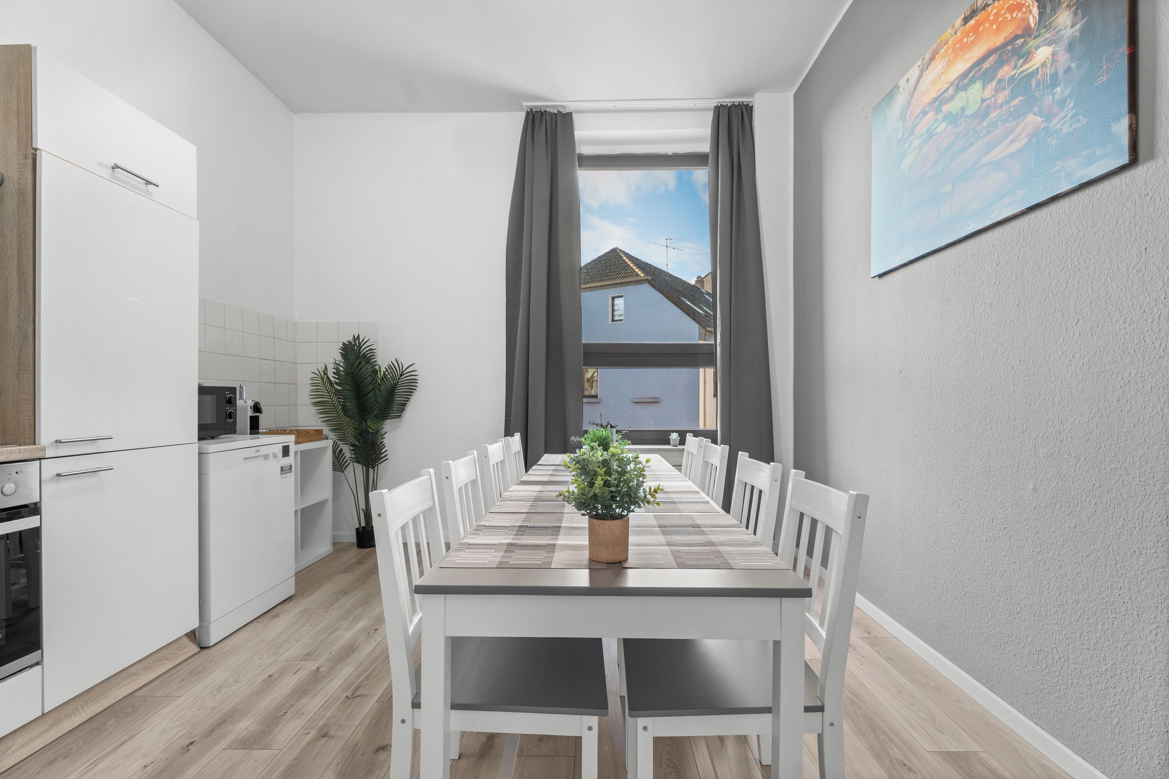 Una cocina moderna con armarios blancos, una mesa de comedor con sillas blancas, una planta en maceta y vistas a una casa a través de una ventana.