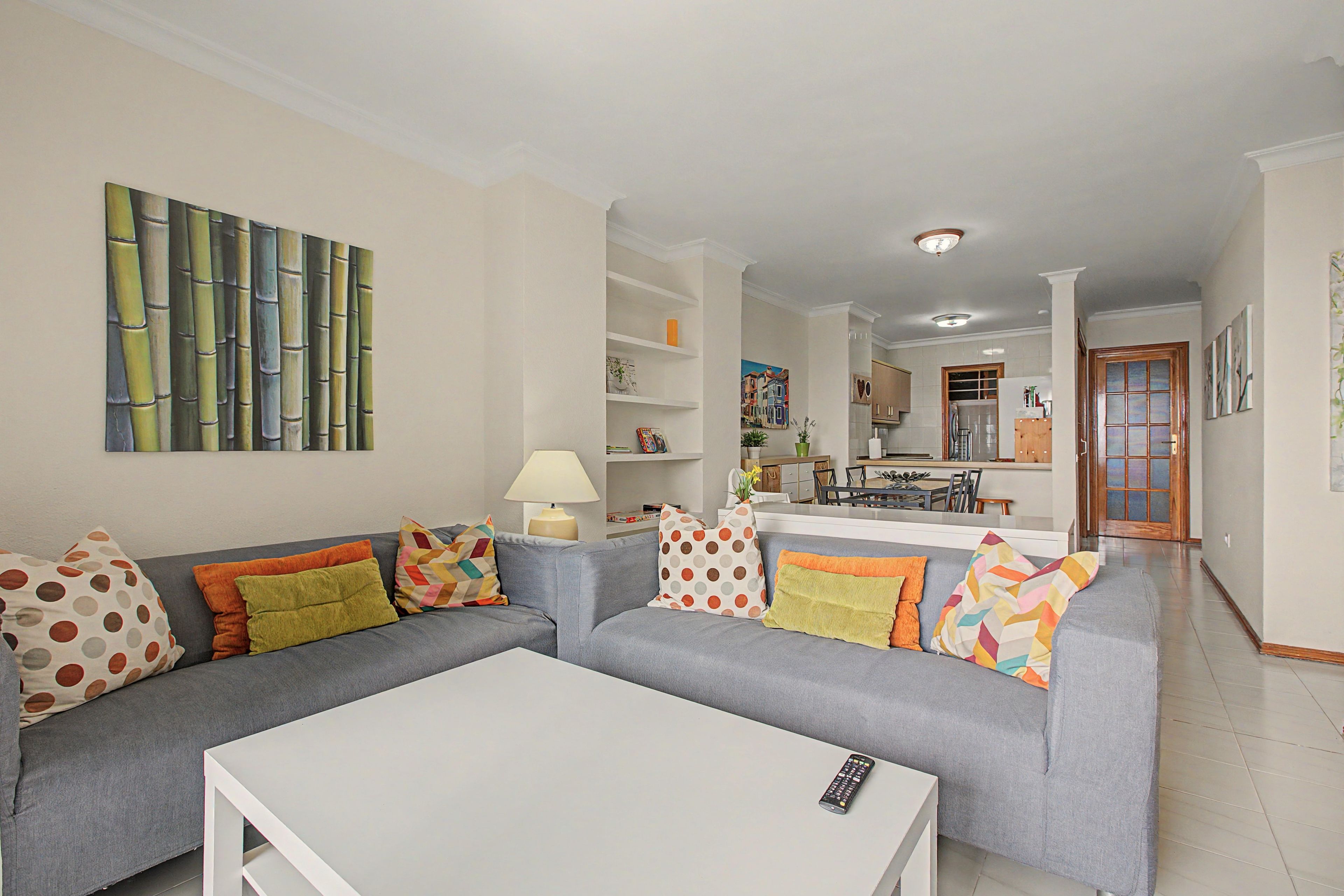 Una sala de estar con un sofá gris, una mesa de centro blanca y almohadones de colores. Hay un cuadro en la pared y una cocina al fondo.