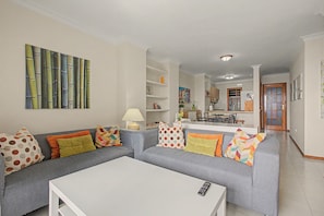 A living room with a grey sofa, white coffee table, and colorful pillows. A painting is on the wall, and there is a kitchen in the background.