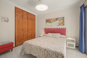 A bedroom with a bed, a nightstand, a red bench, a wooden wardrobe, and a painting on the wall.