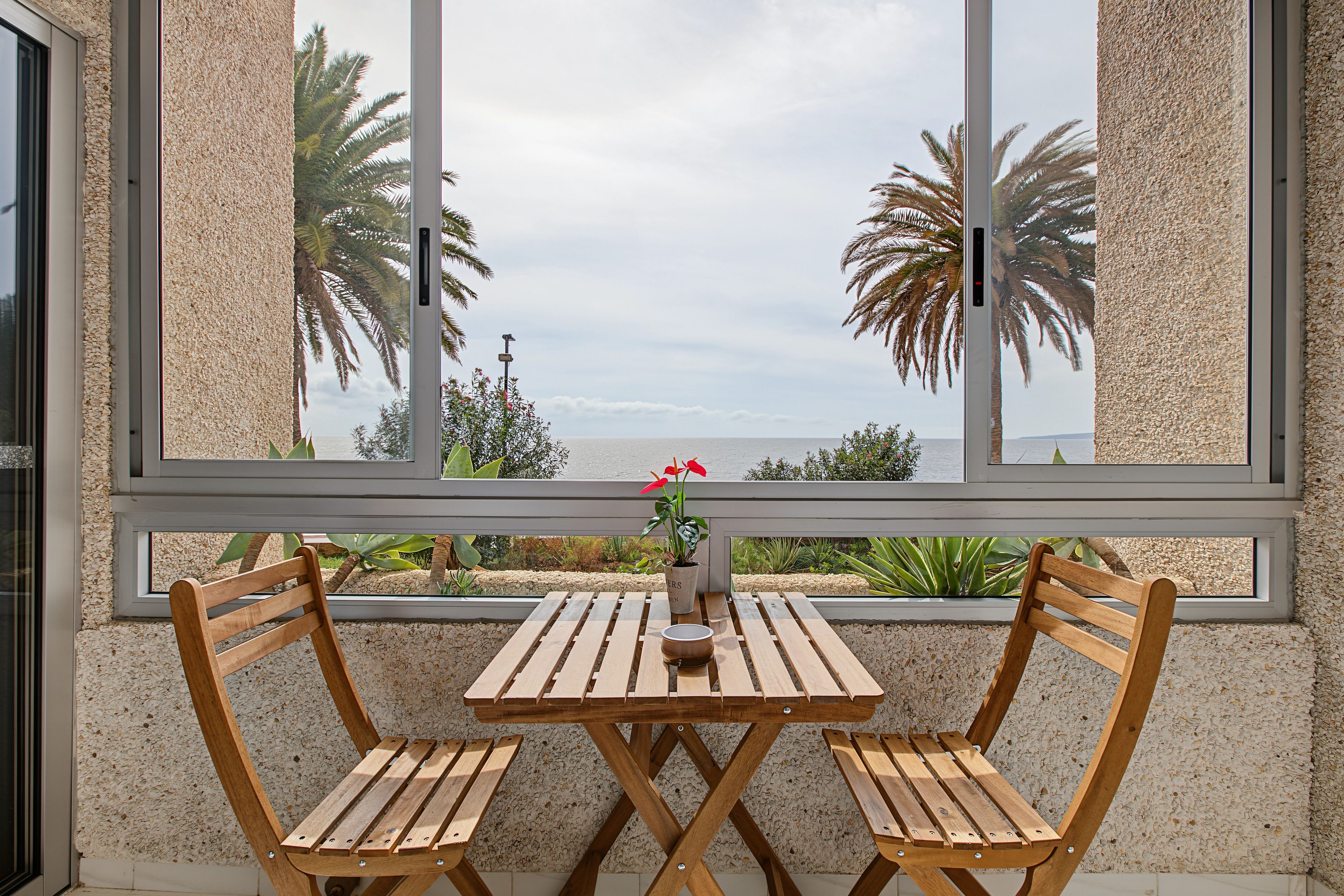 Una mesa de madera y dos sillas frente a una ventana con vista a palmeras y al mar.