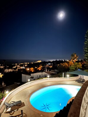 Ein Nachtblick auf einen Poolbereich mit sichelförmigem Mond am Himmel, umgeben von einer Wohngegend mit Häusern und Straßenlaternen.