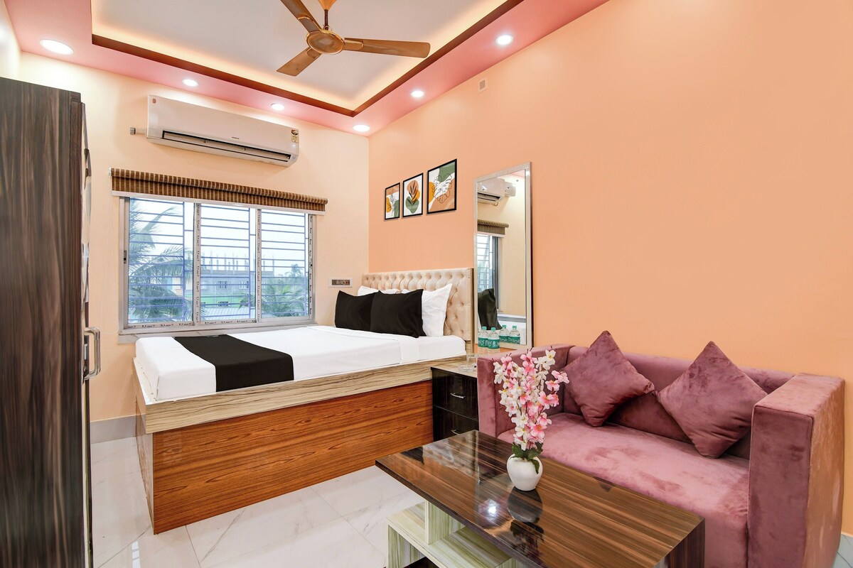 A hotel room with a bed, a sofa, a coffee table, an air conditioning unit, a ceiling fan, a window with blinds, and a mirror.