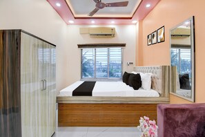 Une chambre à coucher comprenant un lit, une armoire, un appareil de climatisation, un ventilateur de plafond et une fenêtre donnant sur des immeubles.