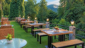 An outdoor dining area with tables and chairs, surrounded by greenery and stone pillars with lights.
