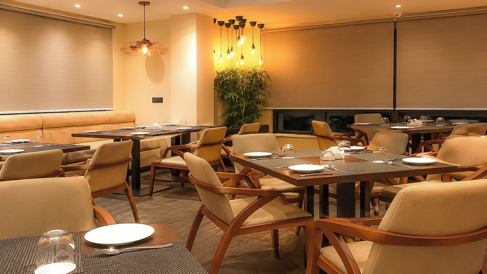 A hotel dining area with multiple tables and chairs, a large plant, and pendant lighting.