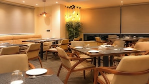 A hotel dining area with multiple tables and chairs, a large plant, and pendant lighting.