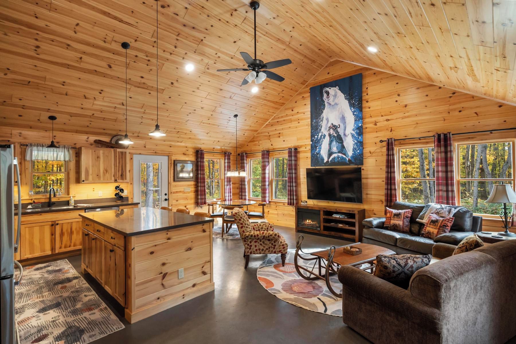 A cabin interior with a kitchen, dining area, and living room, featuring wooden walls and a large window.