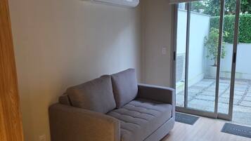 A modern living room with a grey sofa, wooden flooring, and a sliding glass door leading to an outdoor area.