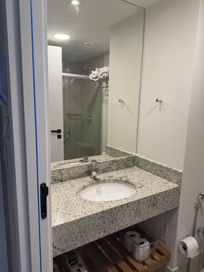 A bathroom with a granite countertop sink, a mirror, a shower, and a toilet paper holder. - New Listing (Niterói)