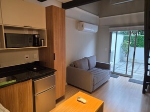 A modern kitchen with a microwave, coffee maker, and a refrigerator. There is a wooden table with a remote control on it. A grey sofa is placed near a sliding glass door leading to a balcony. - New Listing (Niterói)
