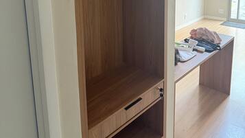 A wooden wardrobe with a hanging rod and a drawer, located in a room with a wooden floor and a window.