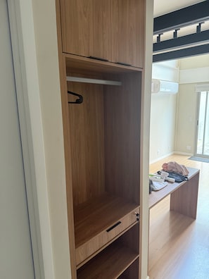 A wooden wardrobe with a hanging rod and a drawer, located in a room with a wooden floor and a window.