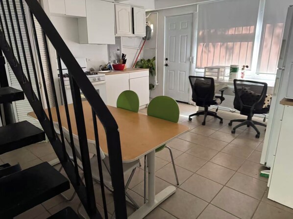 A modern kitchen with white cabinets, a red bowl, and a stove. There is a dining area with a table and two chairs, one green and one black. A staircase is visible in the foreground. - Charming 2-bedroom townhome with AC in vibrant Chicago (Chicago)