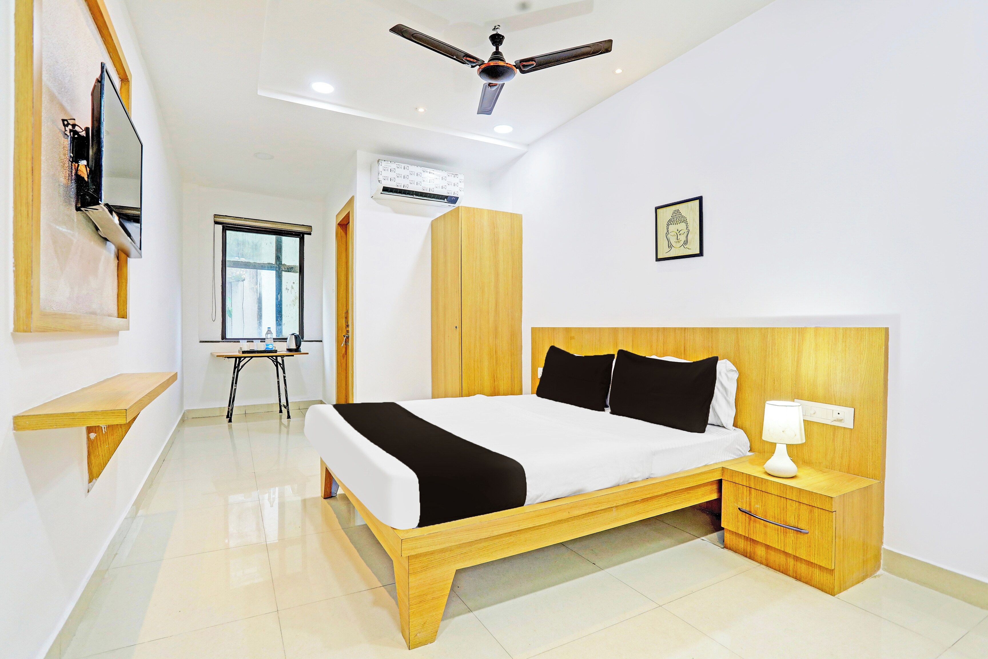 A hotel room with a bed, bedside table, nightstand, ceiling fan, air conditioning unit, window, and a wall-mounted picture.