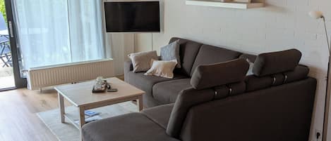 A modern living room with a sectional sofa, a flat-screen TV mounted on the wall, and a wooden ceiling.