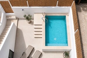 En pool på taget med et klart, blåt vandbassin, omgivet af en hvid facade, en trappe og nogle krukker med planter.