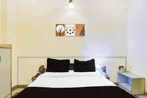 Une chambre d’hôtel avec un lit, deux oreillers noirs, une table de chevet avec une lampe, un vase de fleurs et une œuvre d’art murale au-dessus du lit.