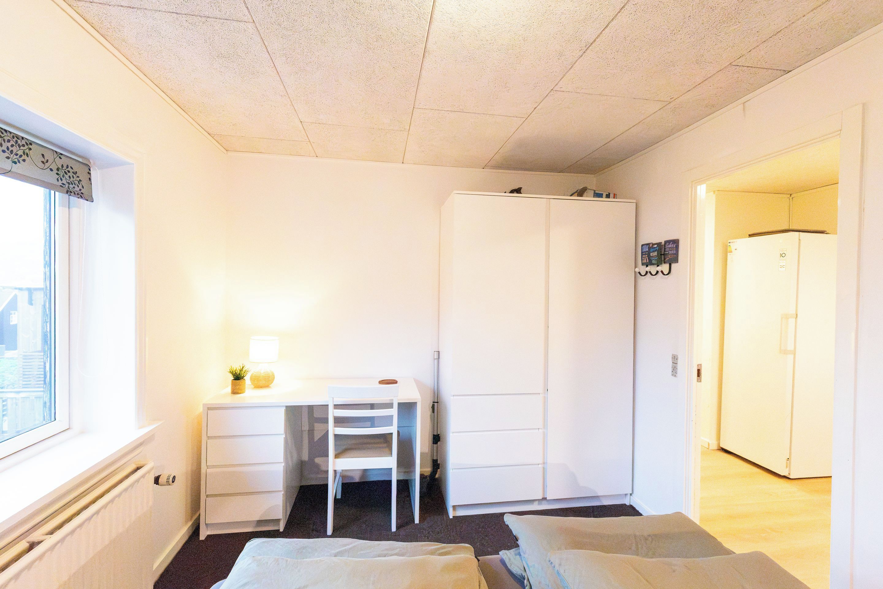 Una habitación pequeña y luminosa con una cama individual, un escritorio con silla, un armario y una puerta que da a otra habitación.