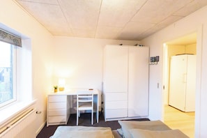 Ett litet, väl upplyst rum med en säng, ett skrivbord med en stol, en garderob och en dörr som leder till ett annat rum.