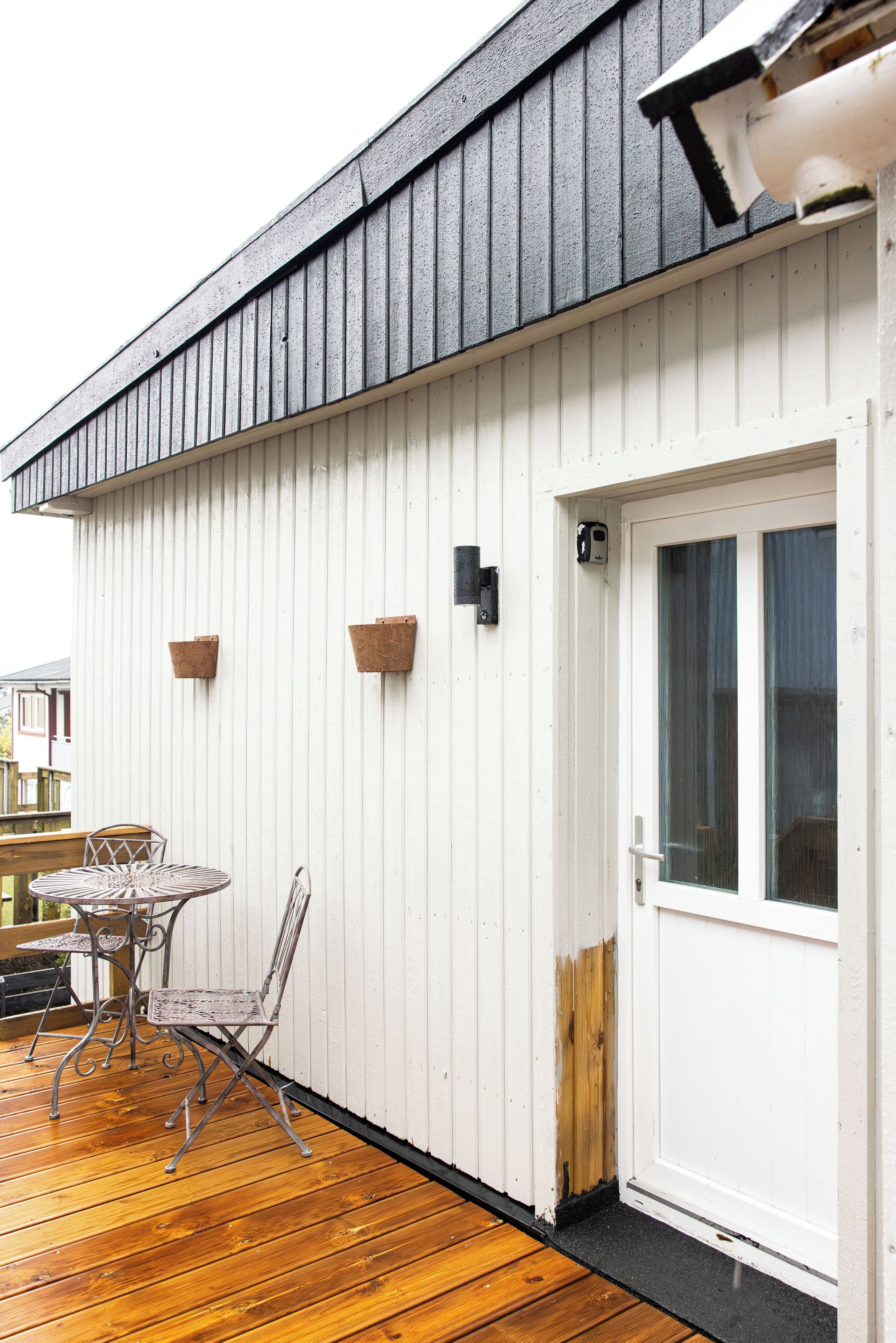 Una terraza de madera con una mesa y sillas de metal, una pared exterior blanca y una puerta con ventanal.