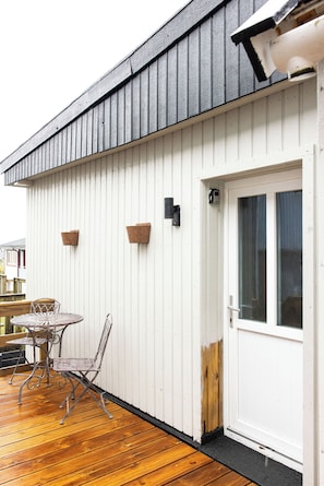 A wooden deck with a metal table and chairs, a white exterior wall, and a door with a window. - Kommunubrekka 8B (Klaksvík)