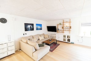 A modern living room with a sofa, coffee table, and a flat-screen TV. There are wall-mounted shelves and a clock on the wall.