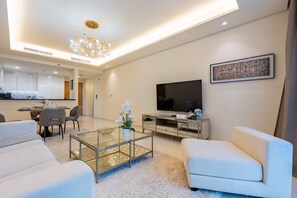 A modern living room with a glass coffee table, white sofas, a flat-screen TV, and a dining area in the background. - High-Floor 1BR, Walking Distance of the Metro and Short Drive from Burj Khalifa (Dubai)