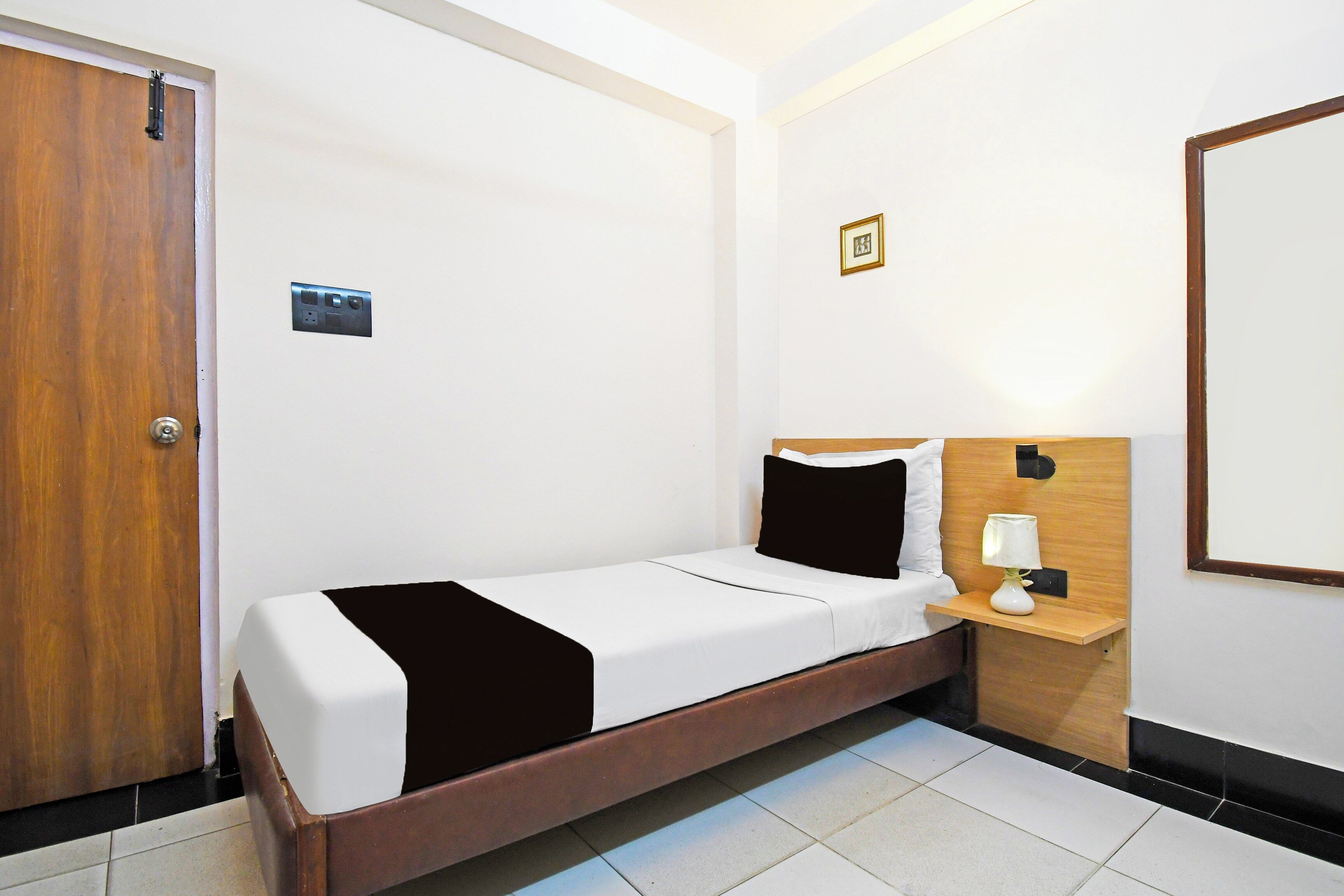 A single bed in a room with a wooden door, a bedside table with a lamp, and a framed picture on the wall.