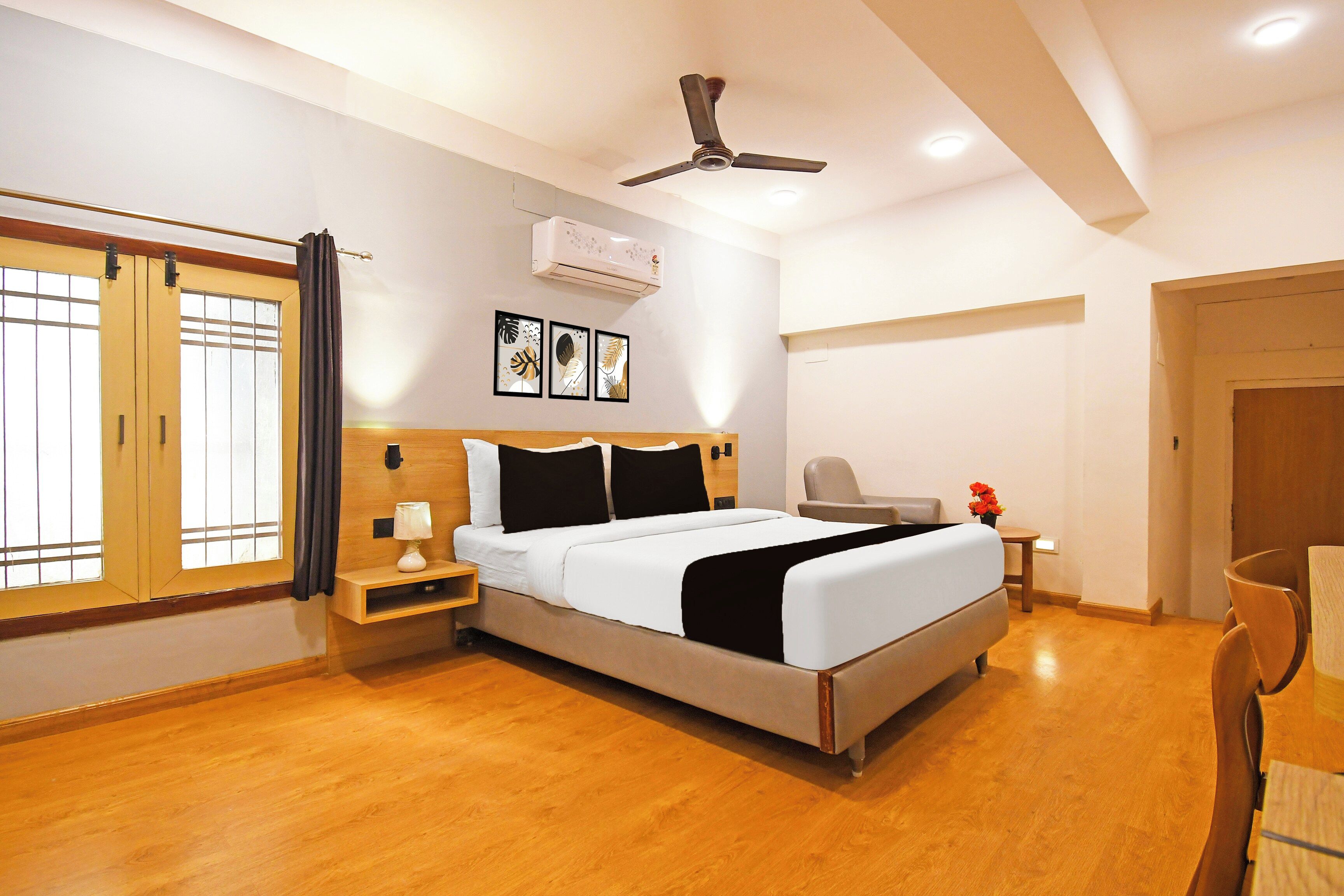 A hotel room with a bed, bedside table, lamp, air conditioning unit, ceiling fan, artwork, and a window with curtains.