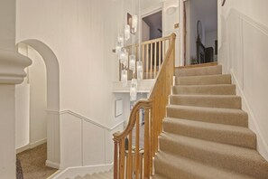 A staircase with beige carpeted steps and wooden railings, leading to a landing with a white wall and a decorative light fixture. - Yule House - North East Escapes (Alnwick)