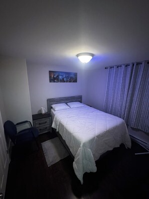 Una camera da letto con un letto, un comodino, una sedia, una finestra con tende e un quadro appeso al muro.