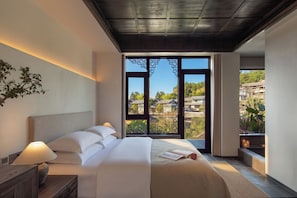 Un dormitorio moderno con una cama grande, una mesita de noche con lámpara y vistas a casas tradicionales a través de un ventanal amplio.