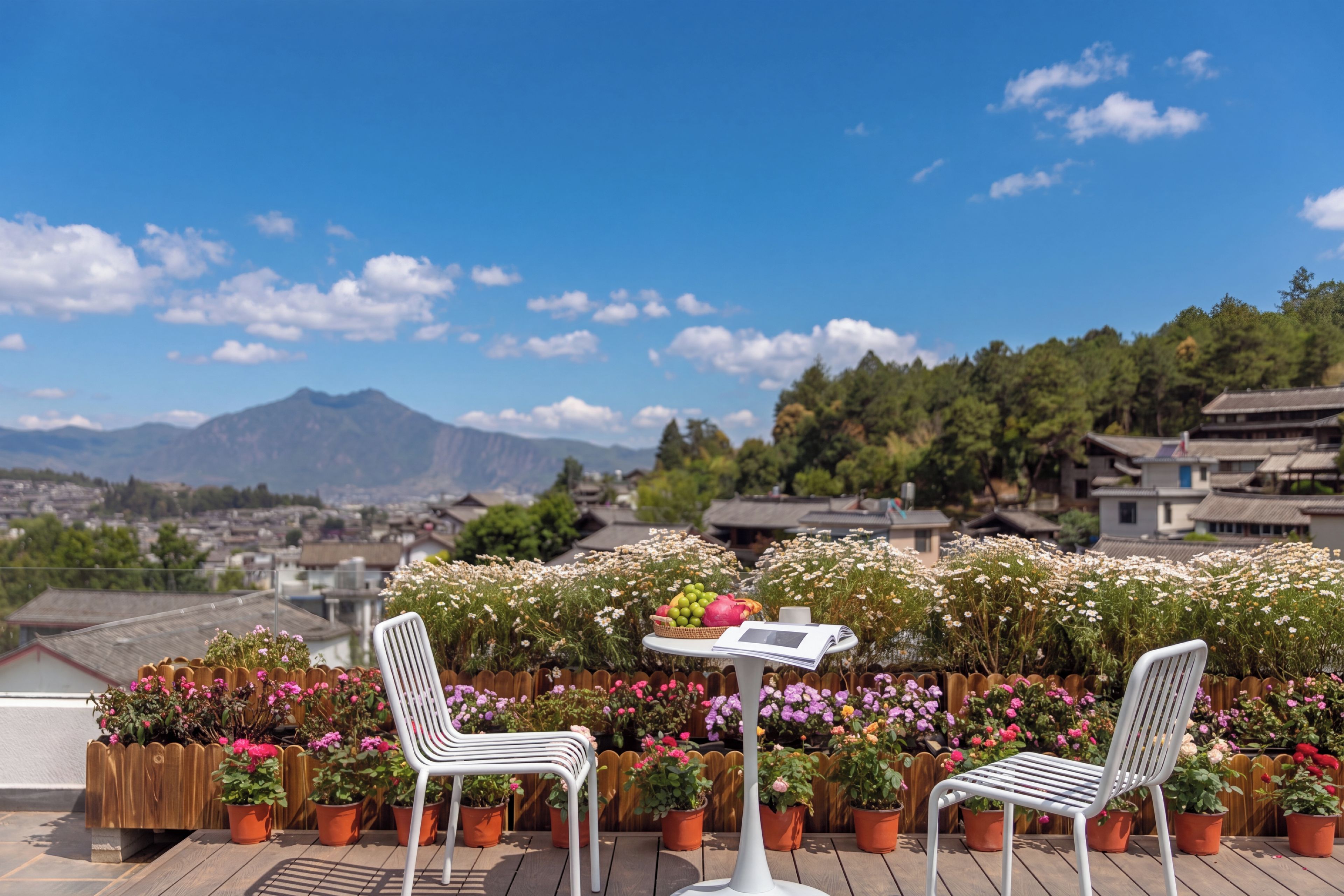 En terrasse med to hvide stole og et bord, omgivet af krukker med planter og blomster, med udsigt over en by og fjerne bjerge.