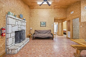 A room with a stone fireplace, a couch, a wooden table, and a ceiling fan. - Riverside Cabins at Cypress Bend #1 @ (Concan)