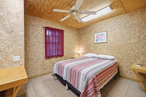 Ein Schlafzimmer mit einem Bett, einem Fenster mit Vorhängen, einem Deckenventilator, einer Nachttischlampe und einem gerahmten Bild an der Wand.