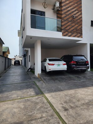 Un edificio moderno con zona de estacionamiento techada, dos coches aparcados y un sendero estrecho que conduce a la entrada.